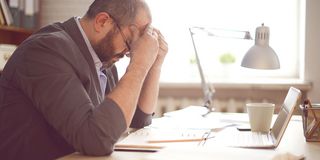 Mann sitzt am Schreibtisch an seinem Laptop und fasst sich konzentriert an den Kopf 