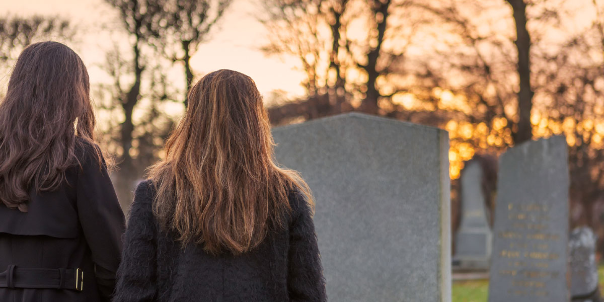 2 Personen die in schwarz gekleidet sind stehen vor einem Grabstein auf einem Friedhof